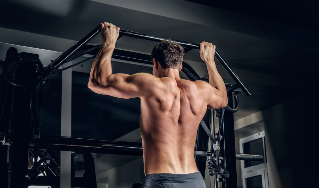 Athletic shirtless man doing pull-ups on a horizontal bar, building upper body strength in the gym.