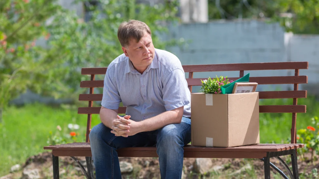 Frustrated man sits on park bench with box after being fired, reflecting on career and life changes